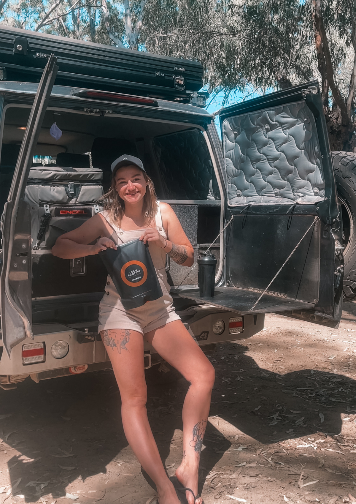 A woman wearing a white cap and beige tank top sits in the open rear of a dark Toyota Landcruiser, holding a black bag of Tropeaka Protein Powder. The vehicle has its doors open, showing a 4wd camping set up inside, with trees and dirt ground in the background.