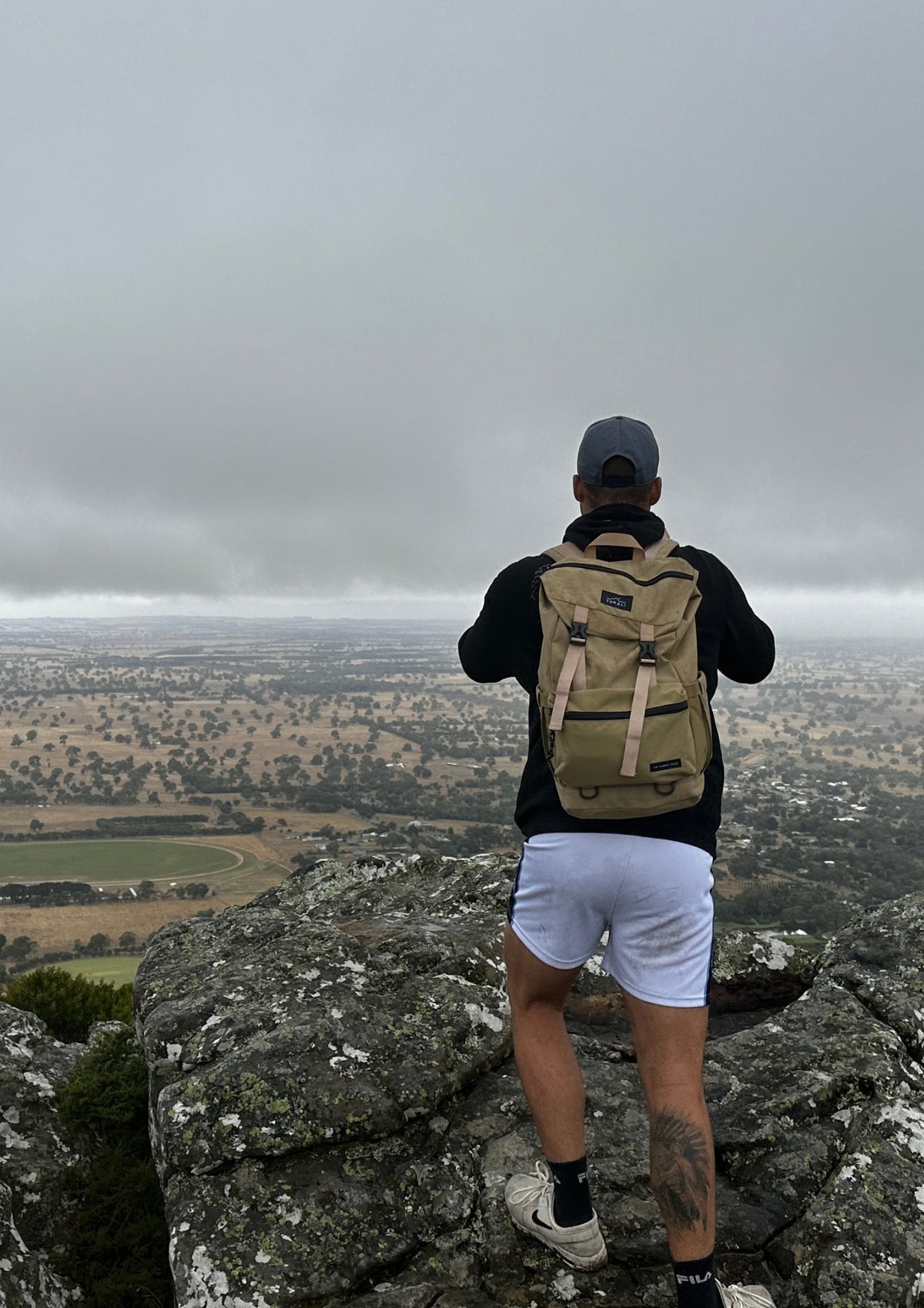 A man stands on a rocky mountain peak with a vast, hazy valley landscape below, viewed from behind as he wears a tan backpack, black long-sleeve shirt, white shorts, and a gray cap.