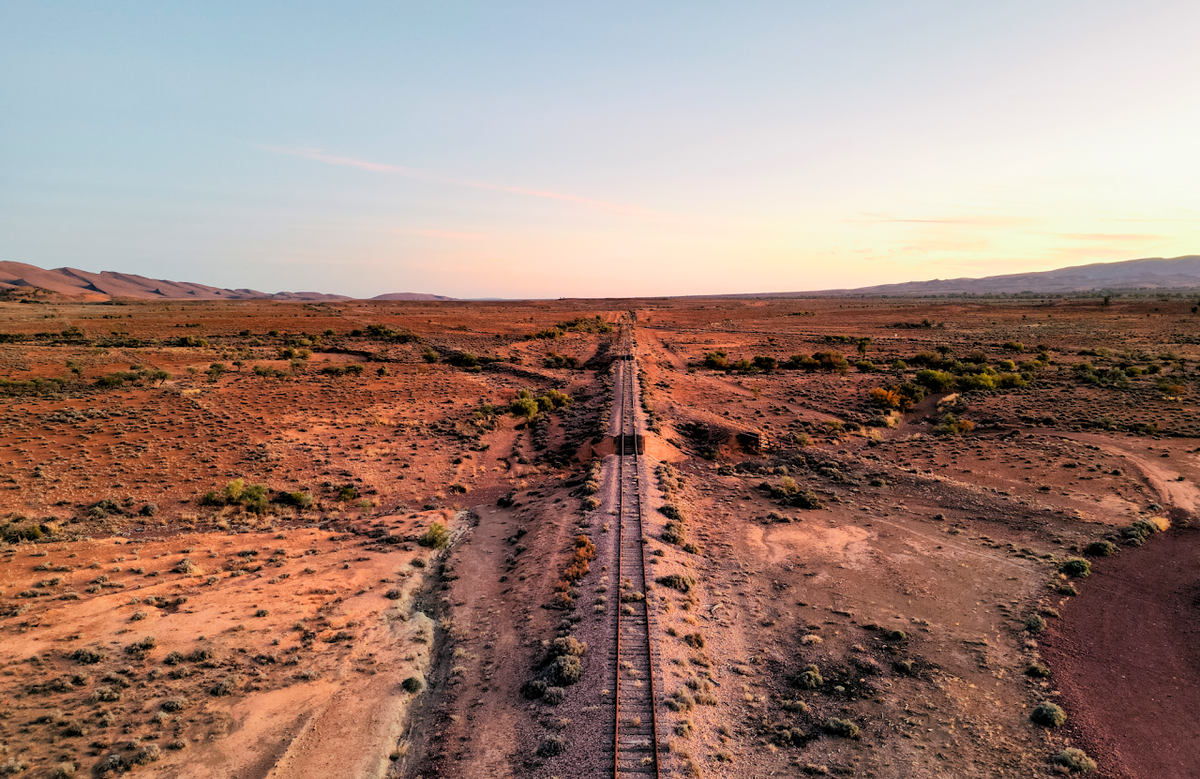 Aerial view of a straight railway track running through a vast red desert landscape under a pale blue and warm sunset sky. This is the old Ghan Railway.