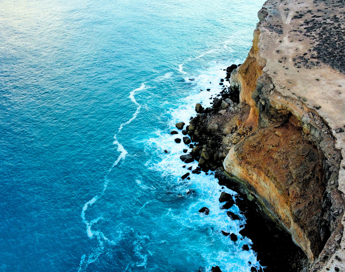 Aerial view of a rugged coastline with bright turquoise ocean water, white surf, dark rocky outcrops, and a tan cliff face on the right. This is the Bunda Cliffs.