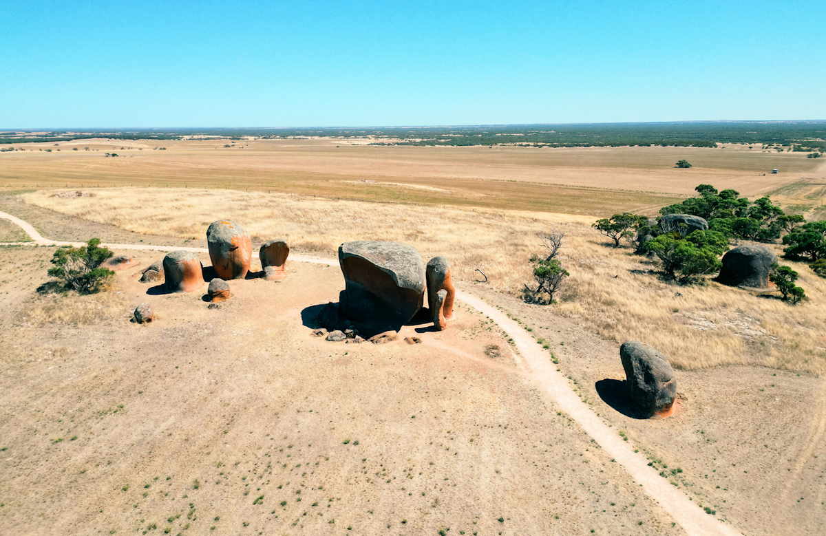 Aerial view of a dry grass landscape with large rock formations, sparse trees, and a dirt path under a clear blue sky. This is Murphys Haystacks.