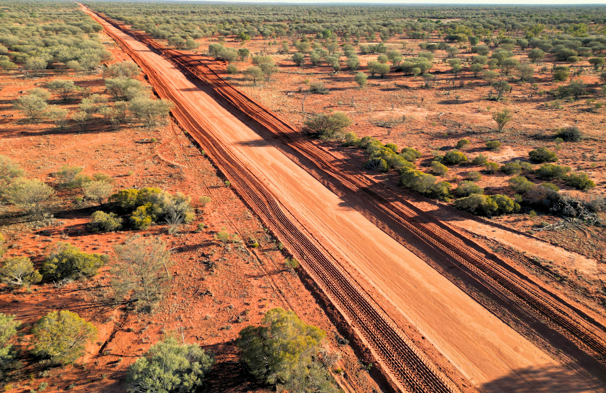 An aerial view of a long dirt road cutting through a red earth landscape dotted with sparse green shrubs and trees.