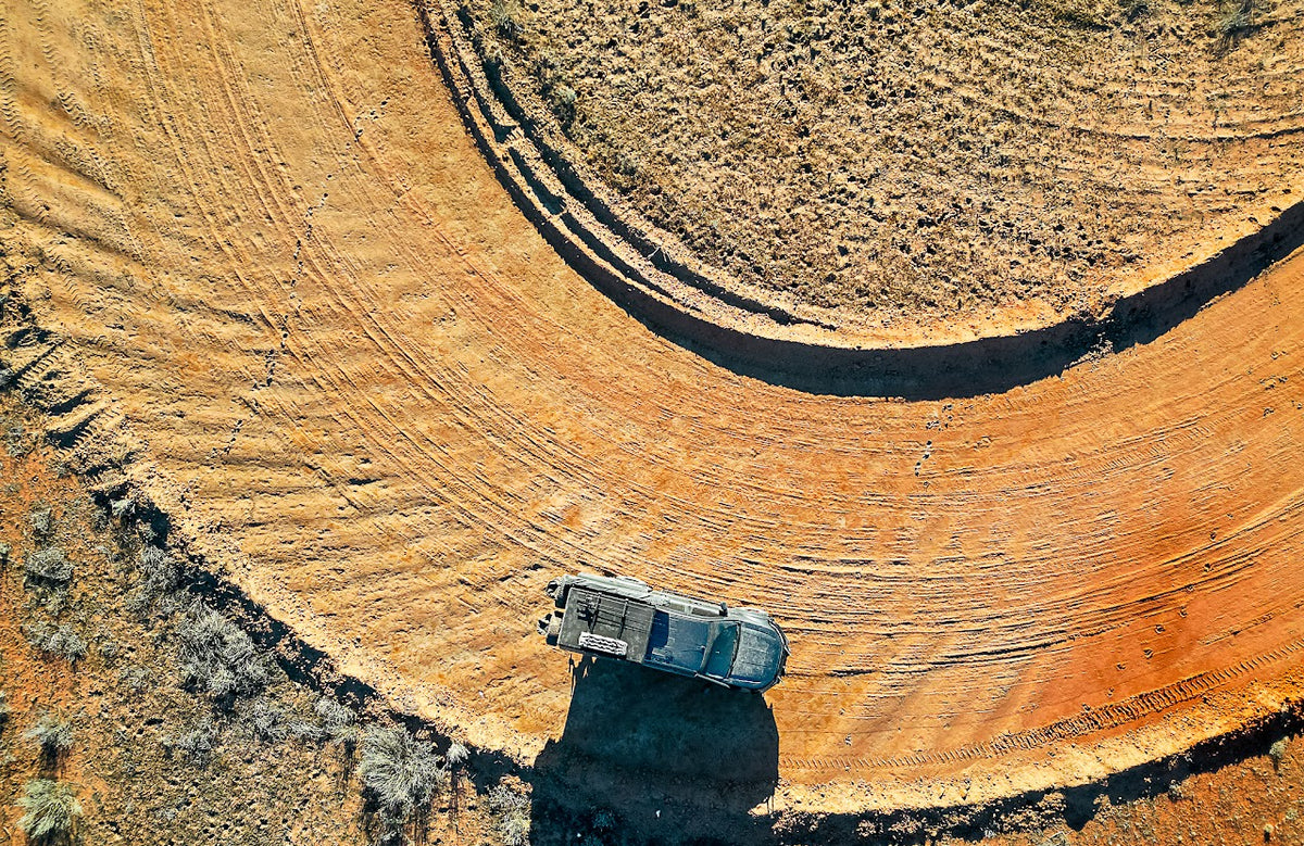 An aerial view of a large curved dirt track or road in a dry, arid landscape, with a small white vehicle positioned near the bottom center on the track.
